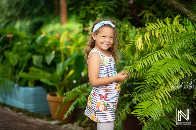 A young girl in a colorful dress stands beside lush green ferns in a bright garden during a sunny day, smiling happily while enjoying the beauty of nature