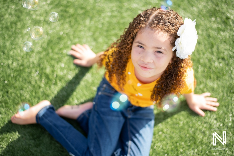 A young girl with curly hair enjoying a sunny day outdoors while surrounded by floating bubbles on green grass at a local park