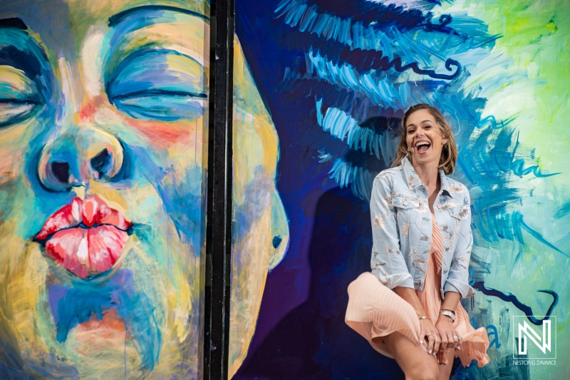 A young woman playfully poses next to a vibrant mural in a colorful urban setting during the day, showcasing artistic street art with a joyful expression