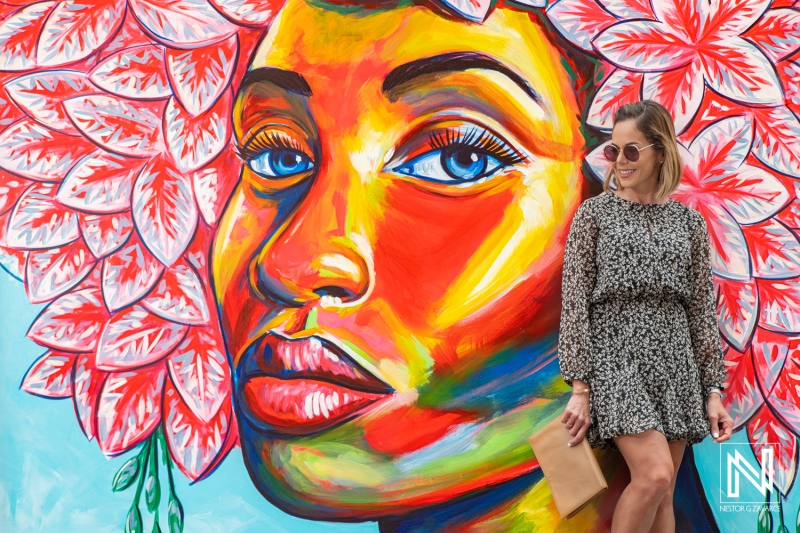 Woman posing against a vibrant mural featuring a colorful portrait of a woman with flower details, showcasing artistic expression in an urban setting during the daytime