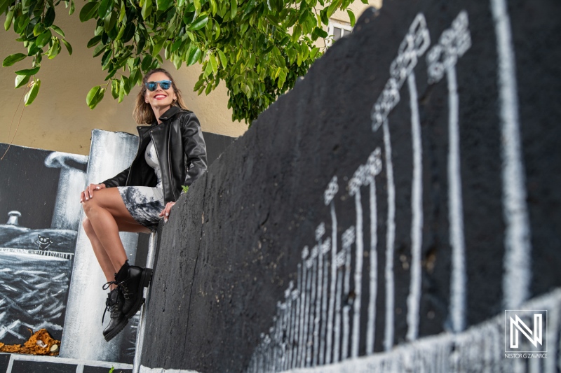 A young woman wearing sunglasses poses confidently against a vibrant mural depicting various artistic elements in an urban setting during daylight