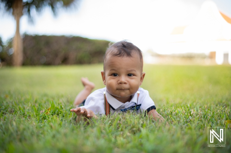 A baby boy in a white shirt with a bow tie lies on green grass, smiling playfully under the warm sunlight in a park during the afternoon