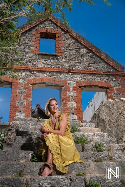 A woman in a yellow dress relaxes on stone steps near an old, partially ruined building under a bright blue sky during the day