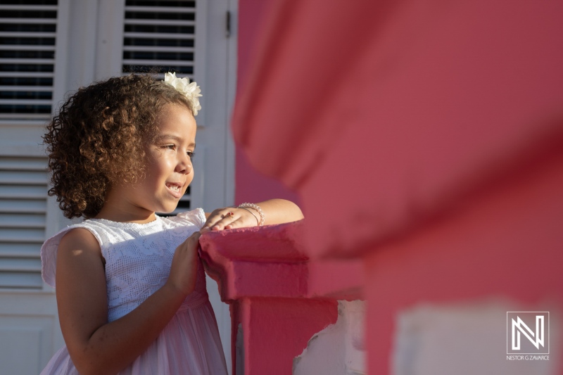 A smiling young girl in a white dress leans against a vibrant pink railing, enjoying a warm afternoon in a charming tropical setting