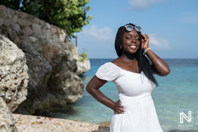 A young woman in a white dress enjoys a sunny day by the turquoise sea, smiling at the camera while standing next to rocky shores in a tropical location