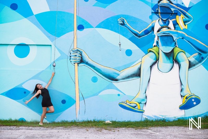 A young woman joyfully poses beside a vibrant mural depicting a colorful fisherman in a scenic outdoor setting during daylight