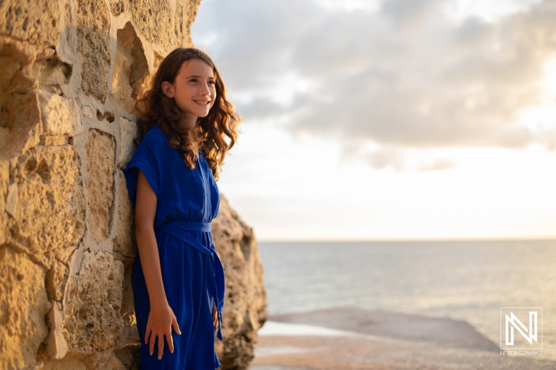 A young girl in a blue dress smiles by the ocean during golden hour, enjoying the serene view at a rocky beach location