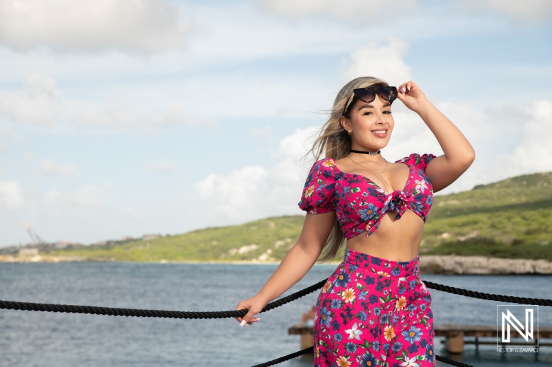 A young woman in a vibrant floral outfit enjoys the sunny weather at a coastal location, playfully adjusting her sunglasses with a scenic view in the background