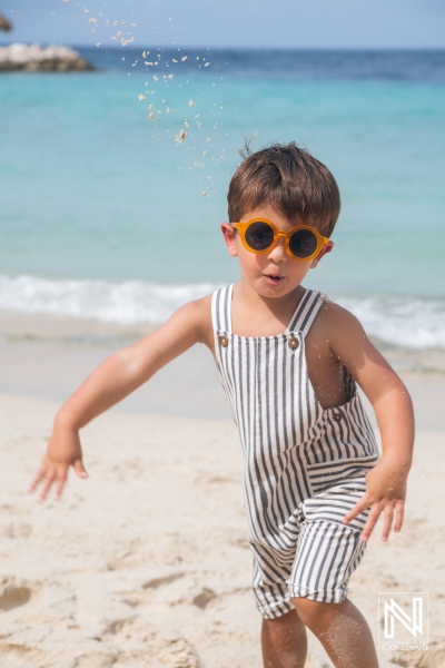 A young boy in striped overalls laughs and plays at the beach while wearing bright orange sunglasses on a sunny day