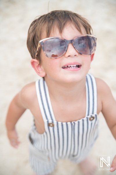 A cheerful young boy in striped overalls and oversized sunglasses enjoying a sunny day at the beach, looking up with a joyful expression during summer adventures