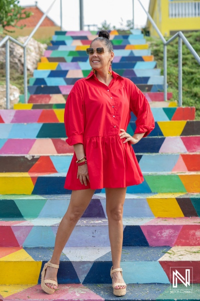 A woman in a bright red dress poses confidently on a colorful staircase in a vibrant outdoor setting during a sunny day