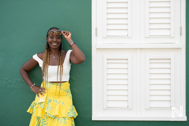 A young woman with braids poses confidently in a yellow skirt and white top against a vibrant green wall on a sunny day in a lively urban setting