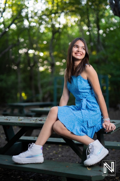 Young girl in a blue dress smiling while sitting on a picnic table in a forested area during daylight hours