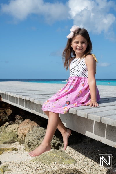 A happy girl in a colorful dress sits on a wooden pier by a beach, enjoying a sunny day with a beautiful blue ocean in the background