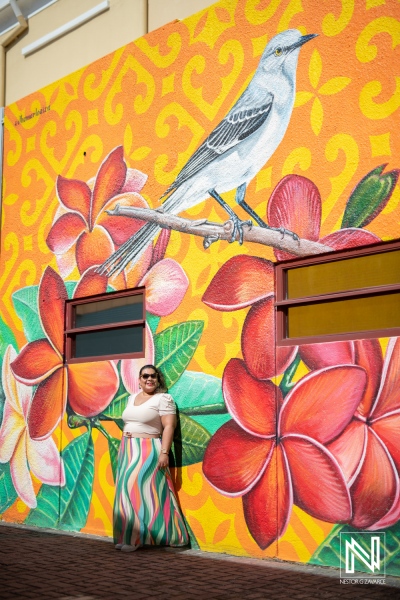 A woman poses next to a vibrant mural of flowers and a bird in a colorful urban setting during a sunny day