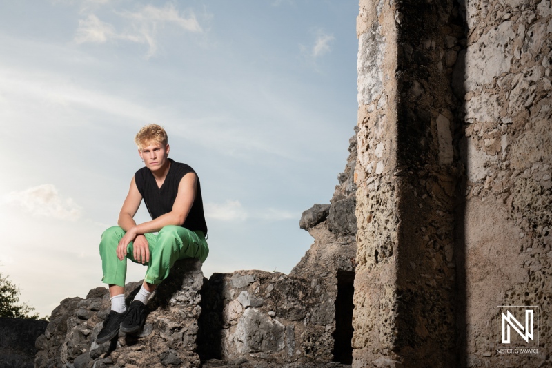 A young man with blonde hair sits on ancient stone ruins wearing green pants and a sleeveless black shirt during a calm sunny day near a historical site