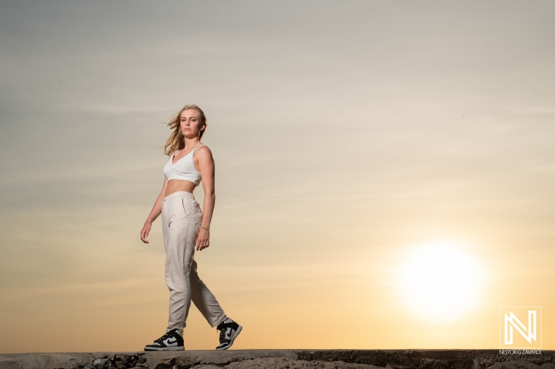 Young woman walking on rocky shore during sunset, wearing casual clothing and sneakers, with a vibrant sky in the background emphasizing the tranquil atmosphere