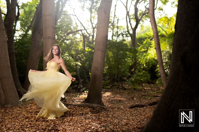 A woman twirls in a flowing yellow gown amidst tall trees and autumn leaves in a serene forest environment during daylight hours