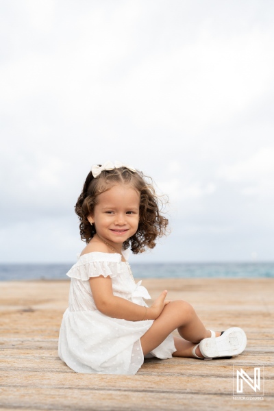 A young girl with curly hair sitting on a wooden dock by the sea, enjoying the gentle breeze and sunshine during a warm afternoon