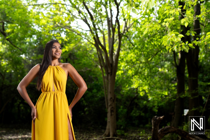 A woman in a yellow dress poses gracefully in a lush green forest during the daytime, highlighting her joy and confidence in the natural surroundings