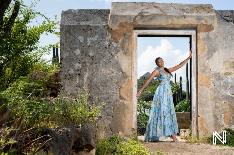 A woman in a flowing blue dress poses at an old stone doorway surrounded by lush greenery and cacti on a sunny day in the countryside