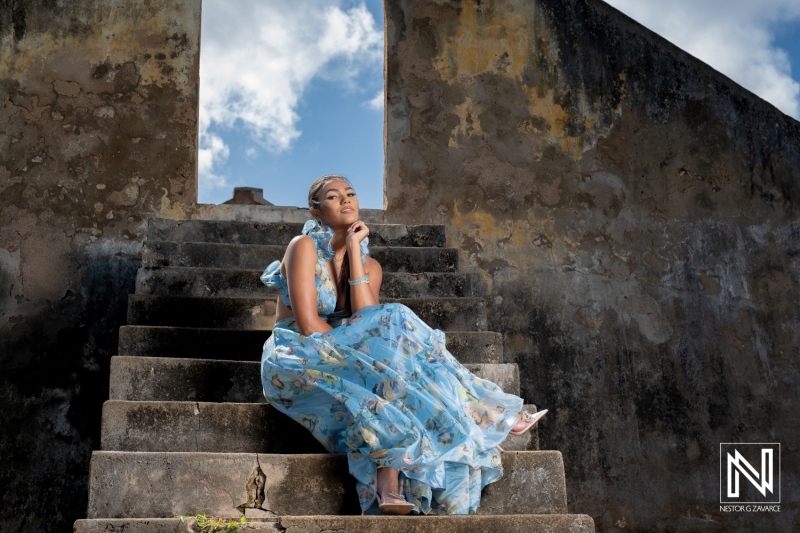 A woman in a flowing blue dress poses gracefully on stone steps under a bright sky in an ancient architectural ruin during a sunny day