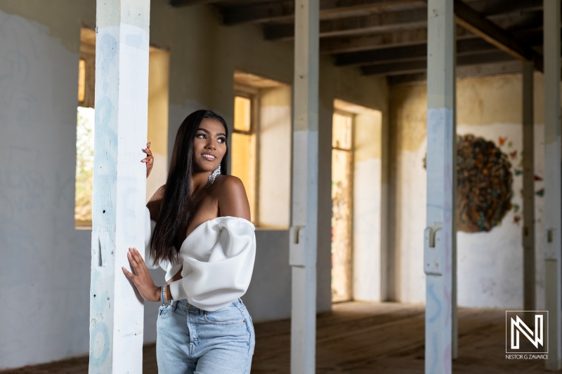 A stylish young woman poses gracefully inside an old building with wooden beams during daylight, showcasing fashion and beauty in a rustic setting