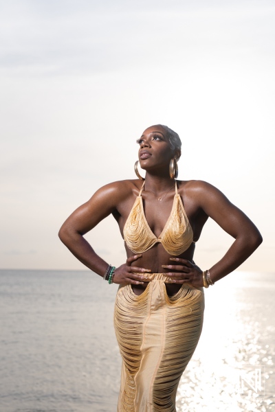 A confident model poses gracefully by the ocean during sunset, showcasing a stunning golden outfit with intricate details while enjoying the serene atmosphere