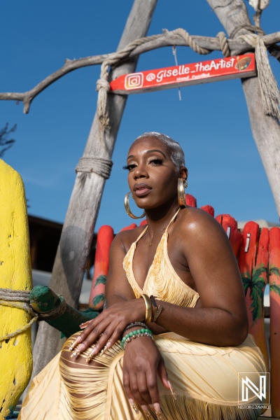 A fashion-forward artist poses gracefully on a colorful wooden chair against a vibrant outdoor backdrop under a clear blue sky during daytime