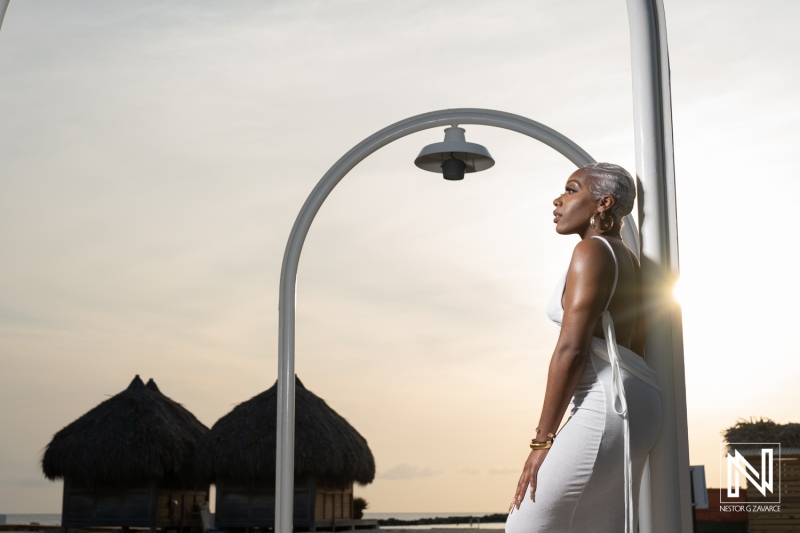 A woman in a white dress stands gracefully under a lamp post at sunset near thatched-roof huts in a tropical destination