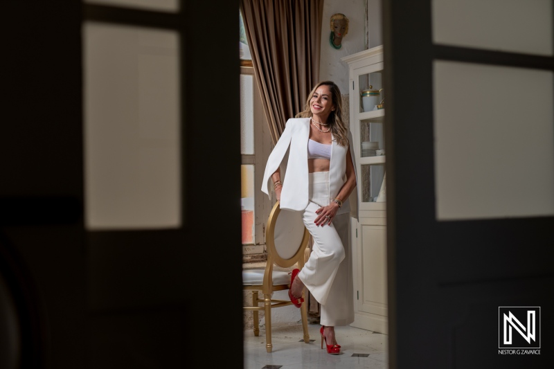 A confident woman in a stylish white suit poses playfully in a well-decorated room with elegant furnishings during daylight hours