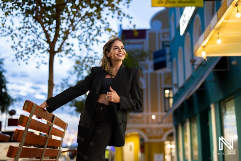 A stylish woman poses confidently on a bench in a vibrant street during sunset, showcasing urban fashion and lively surroundings in a charming neighborhood