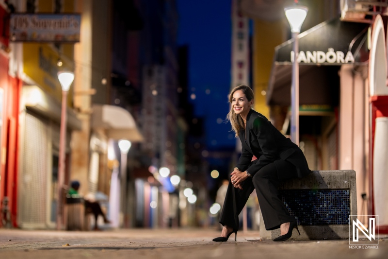 A woman in stylish attire sits confidently on a bench in a vibrant urban alleyway, illuminated by street lights at dusk