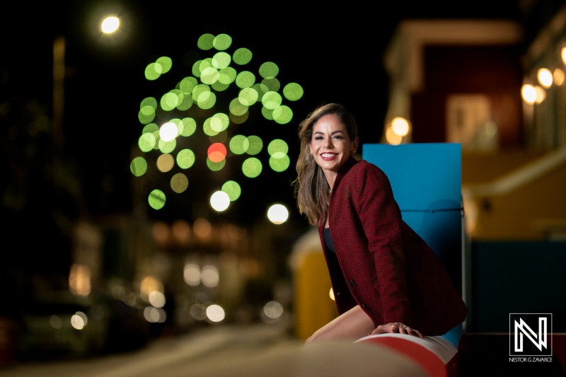 Young woman with a joyful smile sits on a colorful bench in a lively urban setting during a festive night, illuminated by twinkling lights and fireworks in the background