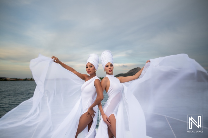 Two elegant models in flowing white attire pose confidently by the water during a sunny day, showcasing a blend of fashion and natural beauty