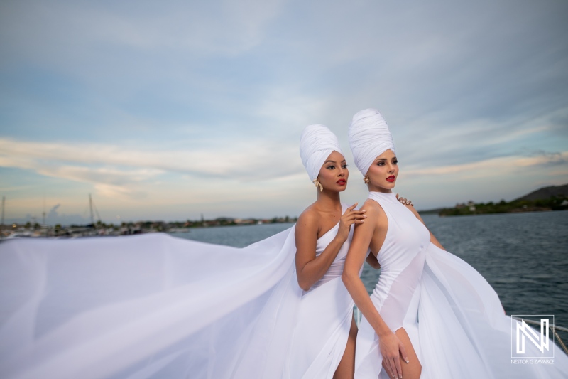 Two models in flowing white gowns pose gracefully near a tranquil waterfront during sunset, showcasing elegance and beauty in a serene setting