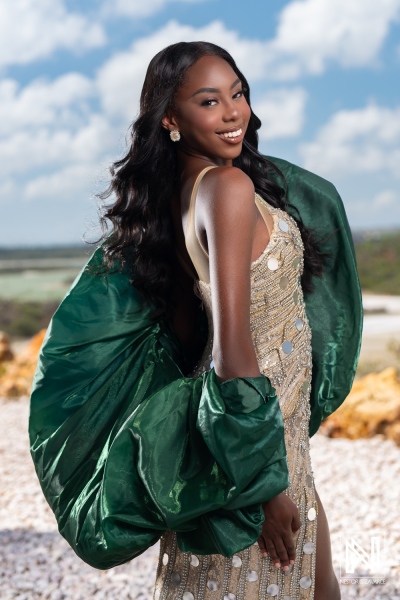 A confident young woman poses outdoors in a shimmering dress with a green cape against a scenic backdrop during the day