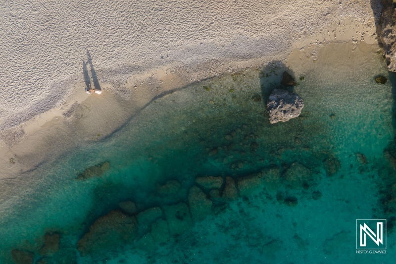 Aerial view of two people walking on a serene beach with clear turquoise water and rocky landscapes during a sunny day in an idyllic coastal location