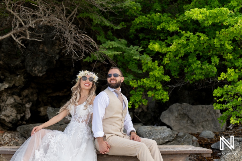 A Joyful Couple Dressed in Wedding Attire Sits Together on a Bench Amidst Lush Greenery, Enjoying a Sunny Outdoor Day