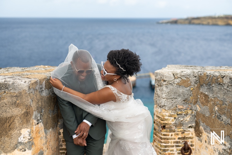 A Joyful Couple Embraces on a Rocky Cliff by the Ocean, Celebrating Their Wedding Day With a Romantic Veil Moment Against a Picturesque Backdrop