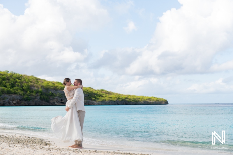 A Couple Shares a Romantic Embrace on a Pristine Beach During Their Wedding Ceremony at Sunset, Surrounded by Lush Greenery and Tranquil Waters