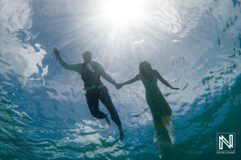 Couple swims together in Curacao waters while holding hands in an underwater scene during bright sunlight