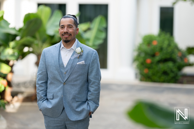 A Groom in a Light Blue Suit With a Boutonniere Stands Confidently Outdoors at a Wedding Venue Surrounded by Lush Greenery on a Sunny Day