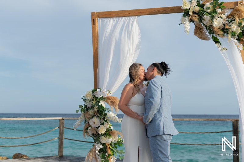 A Couple Exchanges a Kiss During Their Beach Wedding Ceremony at Sunset, Framed by an Elegantly Decorated Wooden Arch With Floral Arrangements