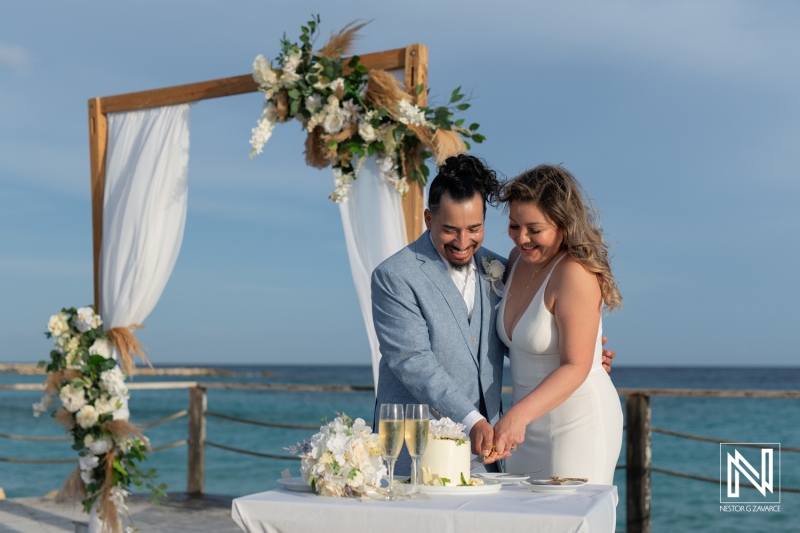 A Couple Celebrates Their Wedding on a Beautiful Beachside Pier, Cutting Their Cake as Waves Gently Lap Against the Shore Under a Clear Blue Sky