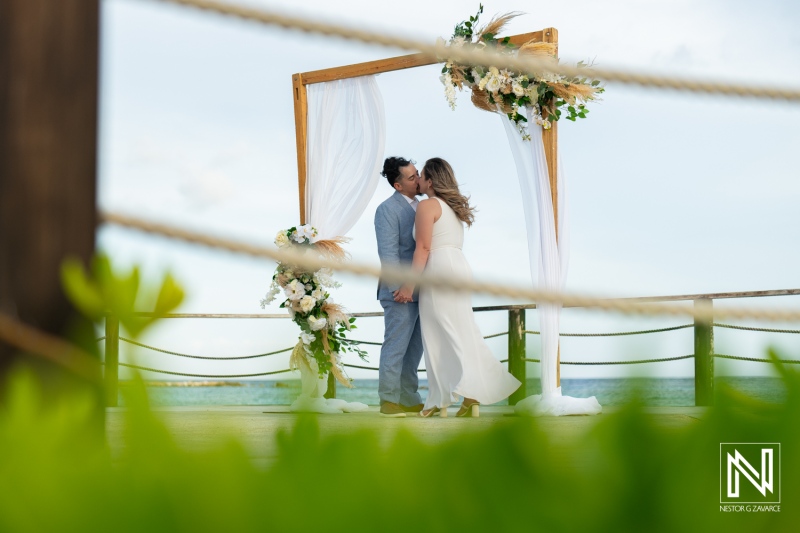 A Couple Shares a Romantic Kiss During Their Beachside Wedding Ceremony With a Decorated Arch and Tropical Scenery, Capturing Their Special Moment at Sunset