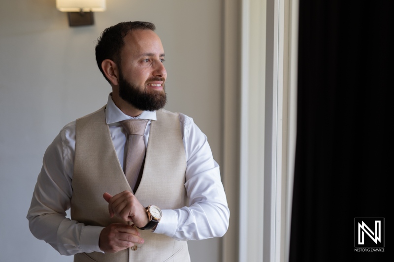 Dapper Man in a Light Vest Adjusting His Watch While Standing Beside a Window in a Well-Lit Room During a Special Occasion