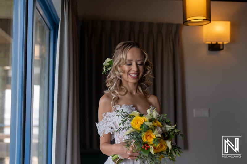 A Joyful Bride in a Floral Wedding Dress Holds a Vibrant Bouquet of Yellow Flowers While Preparing for Her Wedding Ceremony at a Modern Venue in the Late Afternoon Light