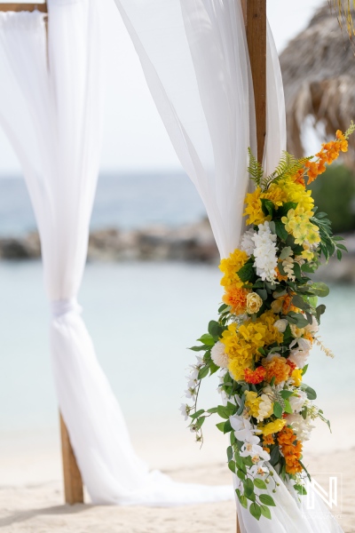 A Beautifully Arranged Floral Arch Draped in White Fabric Stands on a Sandy Beach, Creating a Picturesque Backdrop for a Wedding Ceremony Overlooking the Ocean