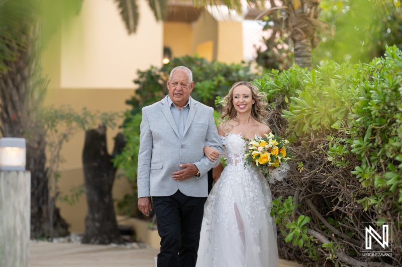 A Bride Walks Down a Sunny Path With Her Father, Carrying a Vibrant Bouquet of Flowers Amidst Lush Greenery at a Tropical Venue